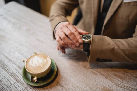 abgeschnittene Ansicht eines Mannes, der eine Armbanduhr in der Nähe einer Tasse Cappuccino mit Latte Art berührt 