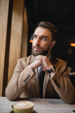 pensive man with beard looking away while sitting near cup of cappuccino in cafe 