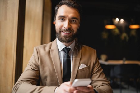 joyful man with beard using smartphone and looking at camera in cafe 