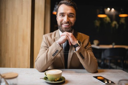 homme heureux avec barbe assis près du smartphone et tasse de cappuccino dans le café 
