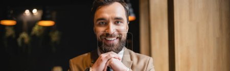 happy man with beard looking at camera while sitting in cafe, banner 