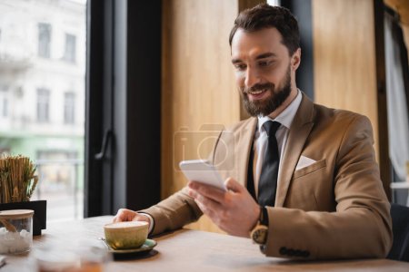 joyful man with beard using smartphone and holding cup of cappuccino in cafe 