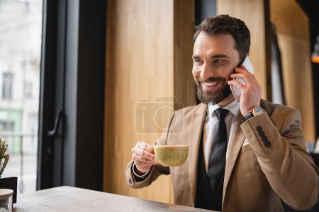 cheerful man with beard talking on smartphone and holding cup of cappuccino in cafe 