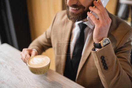 cropped view of cheerful man with beard talking on smartphone and holding cup of cappuccino in cafe 