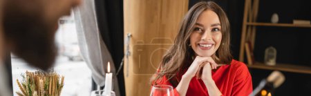 cheerful woman in red dress looking at blurred man on valentines day, banner 