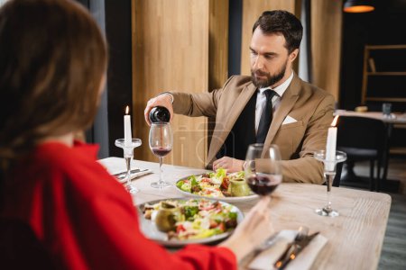 bearded man pouring red wine in glass near girlfriend during dinner on valentines day
