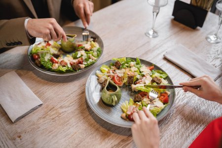 cropped view of couple holding cutlery near salad during lunch in restaurant 