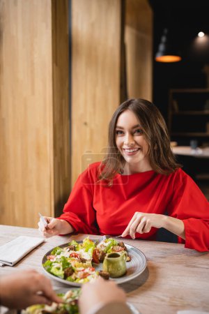 happy woman looking at man in restaurant while holding cutlery near salad in restaurant