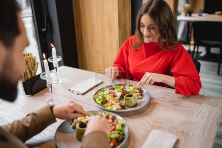 cheerful woman smiling and looking at salad near man in restaurant on valentines day