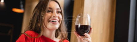 happy young woman holding glass with red wine and smiling in restaurant, banner  