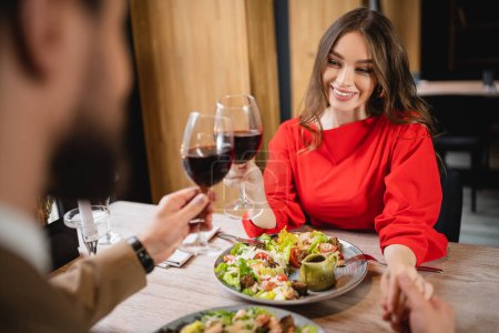 cheerful woman holding hands with boyfriend while clinking glasses of red wine 