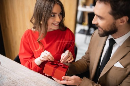 bearded man in formal wear holding present near joyful woman in red dress on valentines day 