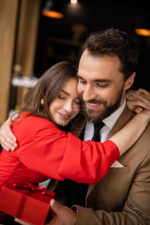 happy woman in red dress hugging bearded boyfriend in formal wear holding present on valentines day 