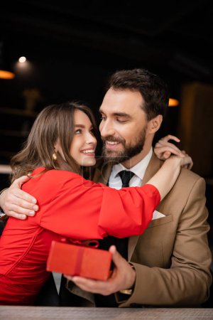 happy woman in red dress hugging bearded man in formal wear holding present on valentines day 