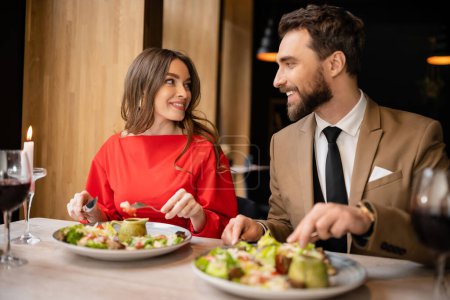fröhliches junges Paar in festlicher Kleidung schaut sich beim Essen während der Feier am Valentinstag an 
