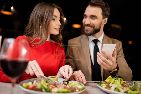 bearded man using smartphone near happy girlfriend in restaurant during celebration on valentines day 