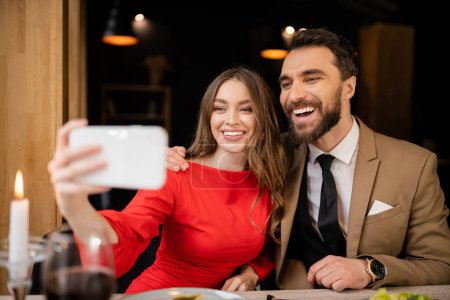 mujer alegre sosteniendo teléfono inteligente mientras toma selfie con el hombre durante la celebración en el día de San Valentín 