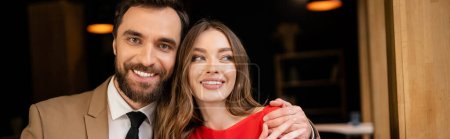 portrait of cheerful and bearded man looking at camera while hugging girlfriend, banner 