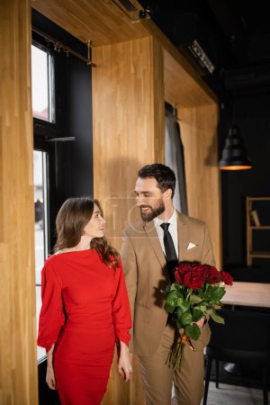barbudo hombre sosteniendo rosas rojas mientras mira alegre novia en vestido en día de San Valentín