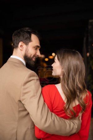 glücklicher bärtiger Mann umarmt fröhliche Freundin im roten Kleid am Valentinstag
