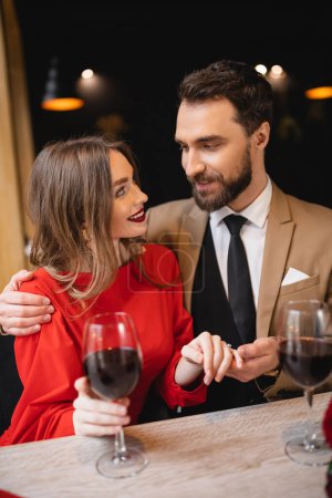 bearded man holding hand of happy woman with engagement ring on finger on valentines day