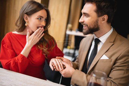 bearded man making proposal and wearing engagement ring on finger of surprised woman on valentines day