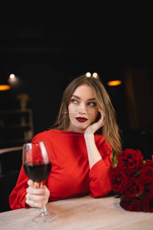 Foto de Joven soñadora sosteniendo copa de vino tinto cerca de rosas en el día de San Valentín - Imagen libre de derechos