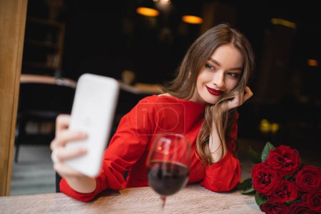 smiling young woman taking selfie near glass of wine and red roses on valentines day