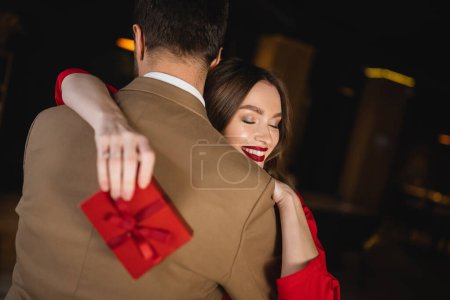 femme gaie avec les yeux fermés tenant présent et étreignant petit ami sur la Saint-Valentin