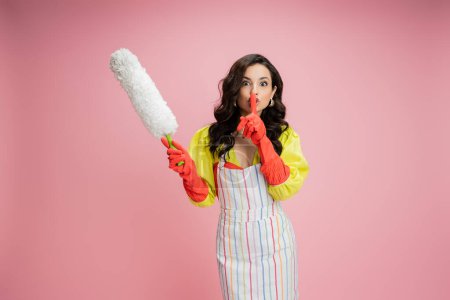brunette woman in yellow blouse and red rubber gloves holding white feather duster and showing hush sign isolated on pink