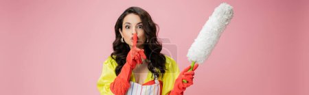 young housewife in red rubber gloves holding white dust brush and showing hush sign isolated on pink, banner