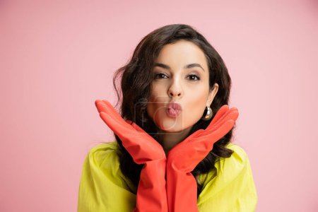 portrait of young brunette housewife in red rubber gloves pouting lips while holding hands near face isolated on pink
