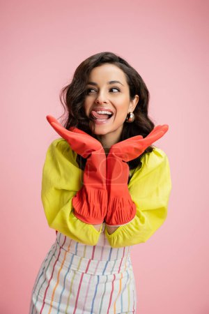 excited housewife in yellow blouse and red rubber gloves holding hands near face and sticking out tongue isolated on pink
