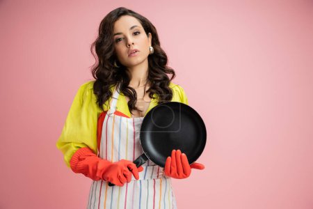 Photo for Pretty brunette housewife in yellow blouse and striped apron holding frying pan and looking at camera isolated on pink - Royalty Free Image
