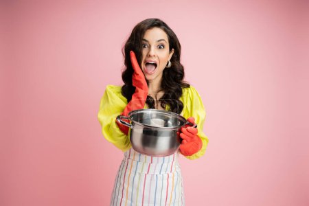 amazed housewife in red rubber gloves holding saucepan and looking at camera isolated on pink