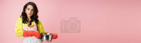 young and pretty housewife holding saucepan and looking at camera isolated on pink, banner