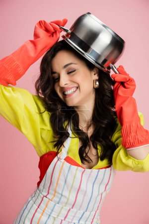 cheerful woman in red rubber gloves having fun while holding saucepan above head isolated on pink