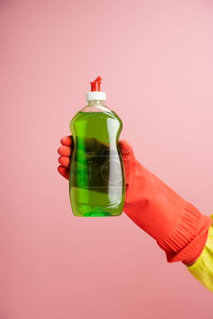 Foto de Vista recortada de ama de casa en guante de goma roja sosteniendo líquido lavavajillas verde aislado en rosa - Imagen libre de derechos