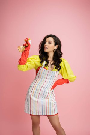 brünette Hausfrau in gelber Bluse und gestreifter Schürze hält Sprühflasche in der Hand und schaut isoliert auf rosa