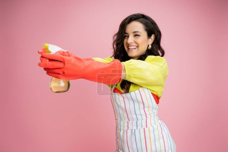 cheerful housewife in striped apron and red rubber gloves having fun and aiming with spray bottle isolated on pink