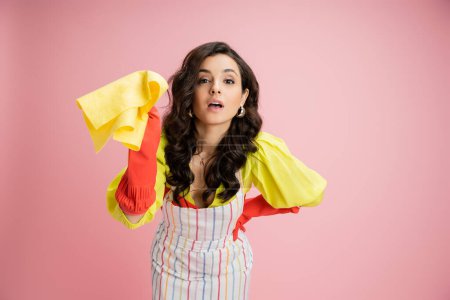 young housewife in striped apron and red rubber gloves holding yellow rag and looking at camera isolated on pink