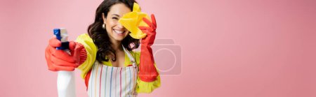 cheerful housewife in red rubber gloves obscuring face with rag and spraying from bottle isolated on pink, banner