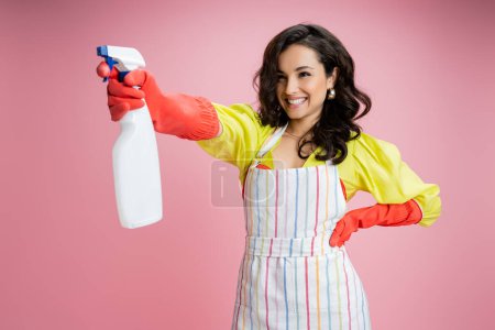 smiling brunette housewife standing with hand on hip and spraying from bottle isolated on pink