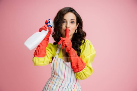 young brunette housewife in striped apron and red rubber gloves holding spray bottle and showing hush sign isolated on pink