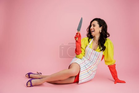 glückliche Hausfrau in gestreifter Schürze und Sandalen, die ein Küchenmesser hält, während sie auf rosa Hintergrund sitzt