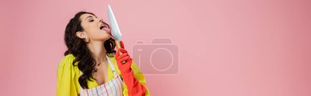 Photo for Pretty brunette housewife in yellow blouse and red rubber glove licking kitchen knife isolated on pink, banner - Royalty Free Image
