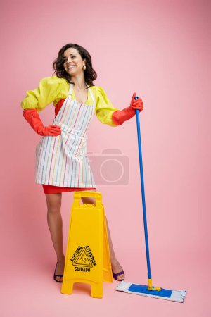 full length of joyful housewife in striped apron standing with mop near wet floor sign and looking away on pink 