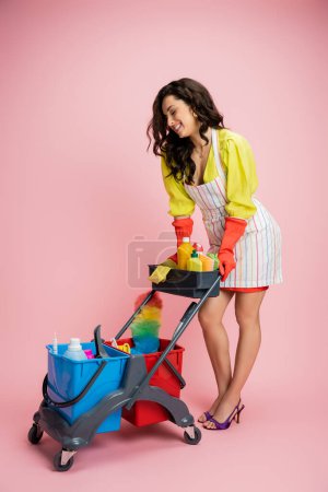 full length of brunette woman in yellow blouse and striped apron smiling near cart with cleaning supplies on pink 