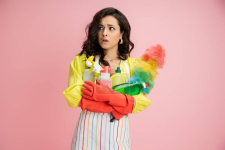 worried housewife in red rubber gloves looking away while holding different detergents isolated on pink