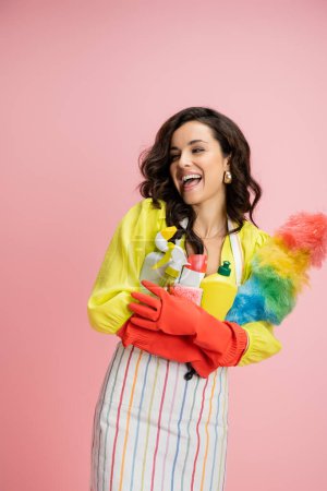 laughing brunette woman in red rubber gloves holding colorful feather duster and detergents isolated on pink